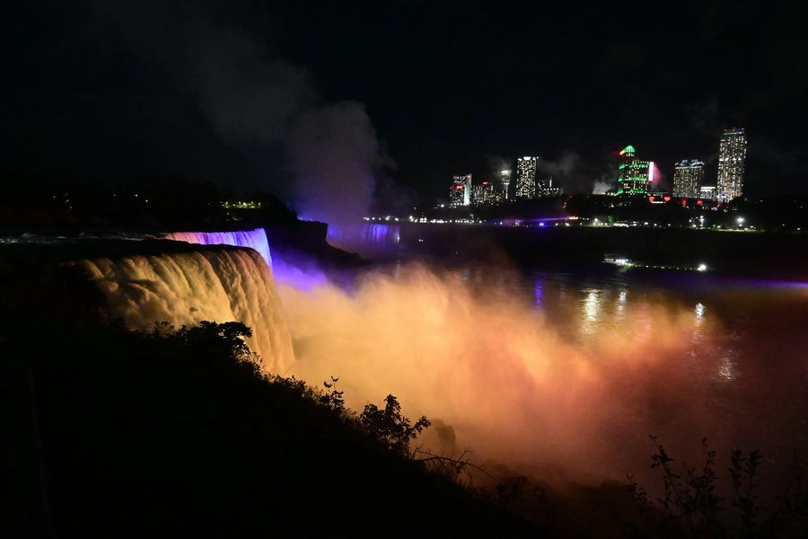 Niagara Falls lit purple and amber at night, mist rising in front of the far skyline.